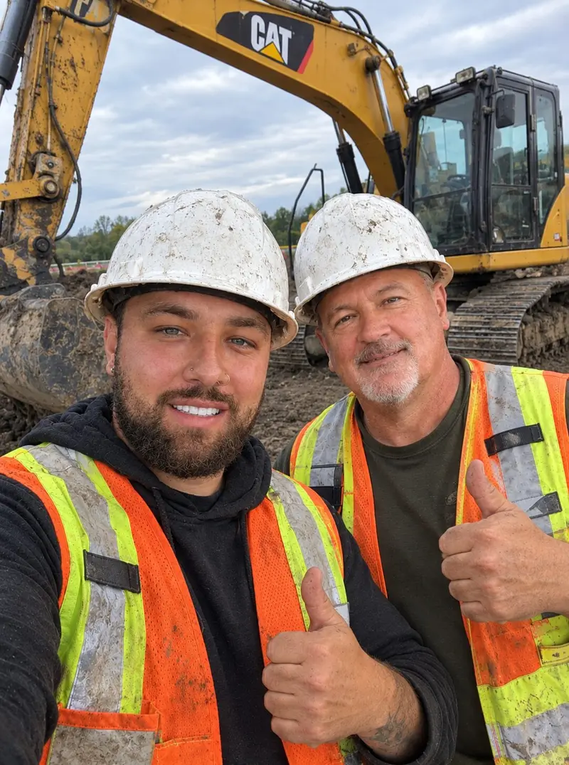 Père et fils de l’équipe AJF Excavation, entreprise d’excavation de Lachine, casques et dossards, pouce levé devant une pelle mécanique